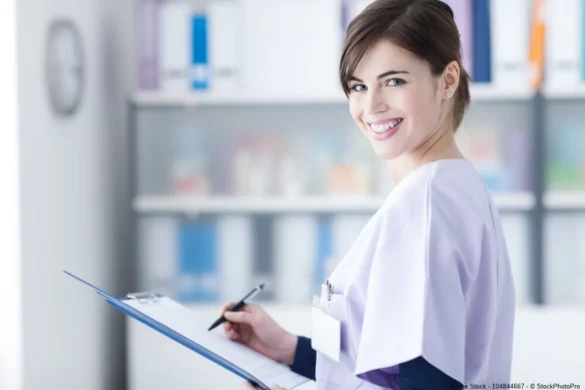 Eine lächelnde Pflegefachkraft mit Klemmbrett und Stift steht in einer medizinischen Einrichtung und symbolisiert Karriere im Gesundheitsbereich.